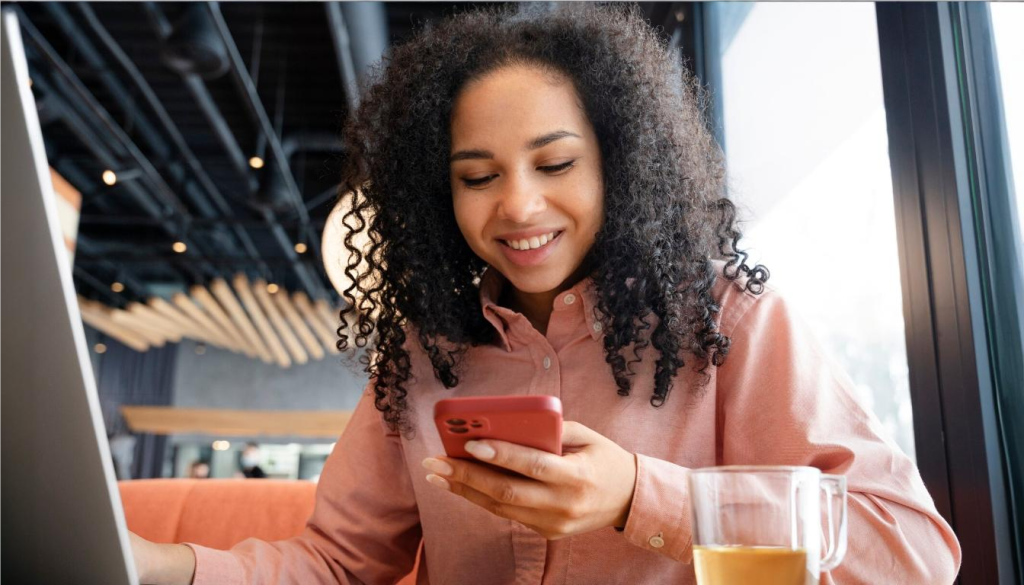 A smiling woman with curly hair sits in a modern café, looking at her smartphone while working on a laptop. A glass mug with tea sits beside her. The image illustrates the flexibility and connectivity offered by unified communications solutions, enabling seamless interaction across multiple devices in remote or hybrid work environments.